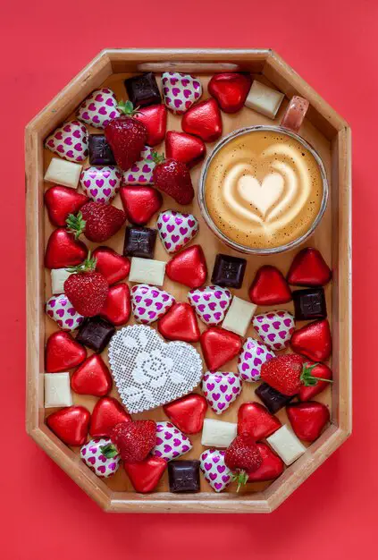 heart-shaped-candies-dark-white-chocolate-strawberries-cup-latte-coffee-charcuterie-board-red-background-close-up-top-view